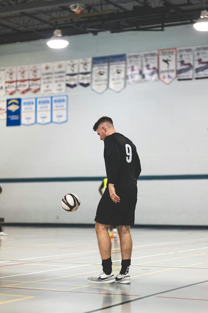 Young soccer player in black jersey with number 9 practices ball control inside a sports hall.